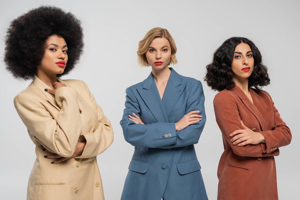 three confident multiracial girlfriends in multicolored suits standing with folded arms on grey - Photo, Image