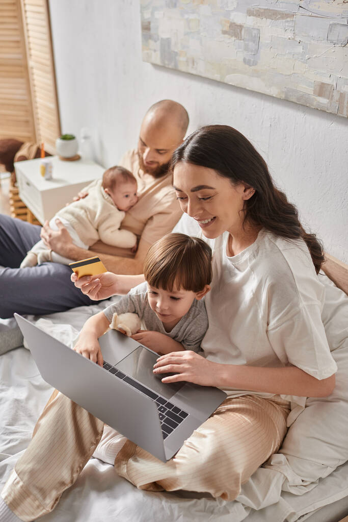 centrarse en la madre con su hijo pequeño con el ordenador portátil y la tarjeta de crédito al lado de marido borroso y recién nacido - Foto, imagen