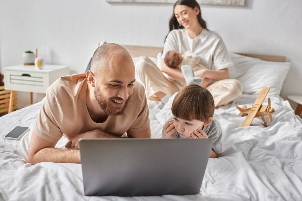 Skup się na wesołym mężczyźnie oglądającym film na laptopie ze swoim synem obok jego rozmytej żony trzymającej dziecko - Zdjęcie, obraz