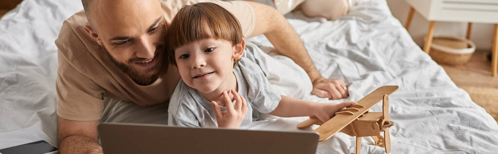 entzückender kleiner Junge mit Holzflugzeug in der Hand und Blick auf Laptop mit seinem bärtigen Vater, Banner - Foto, Bild