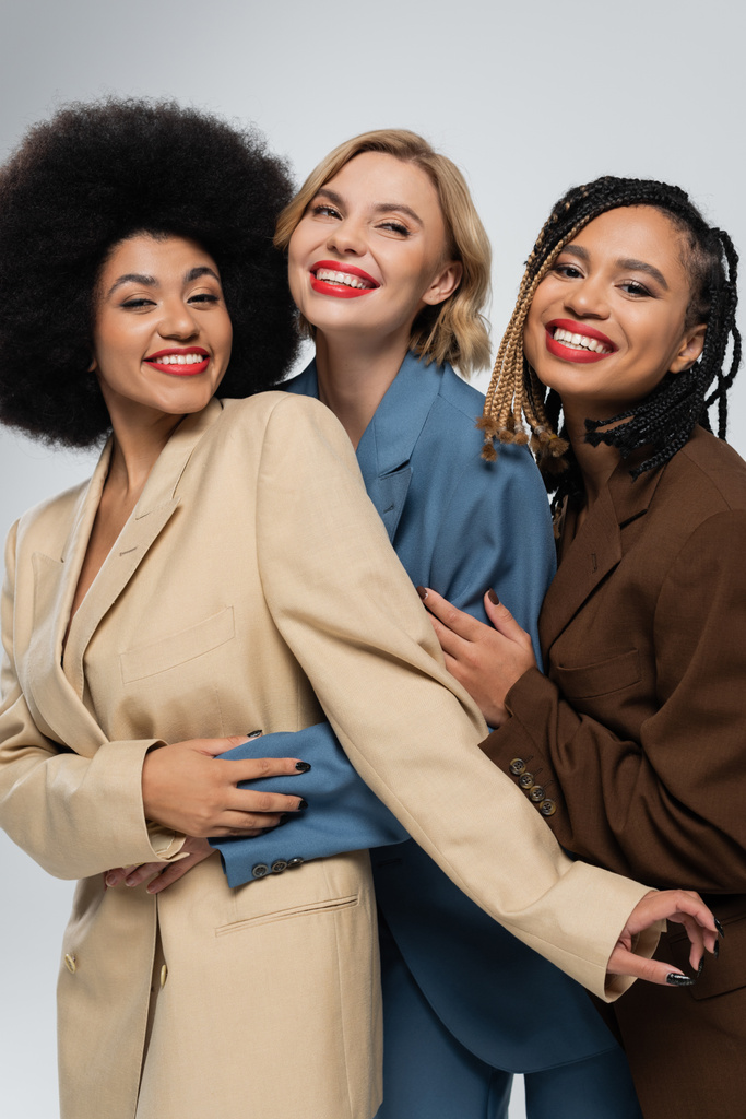 happy multiracial girlfriends in multicolored suits embracing on grey, togetherness and style - Photo, Image