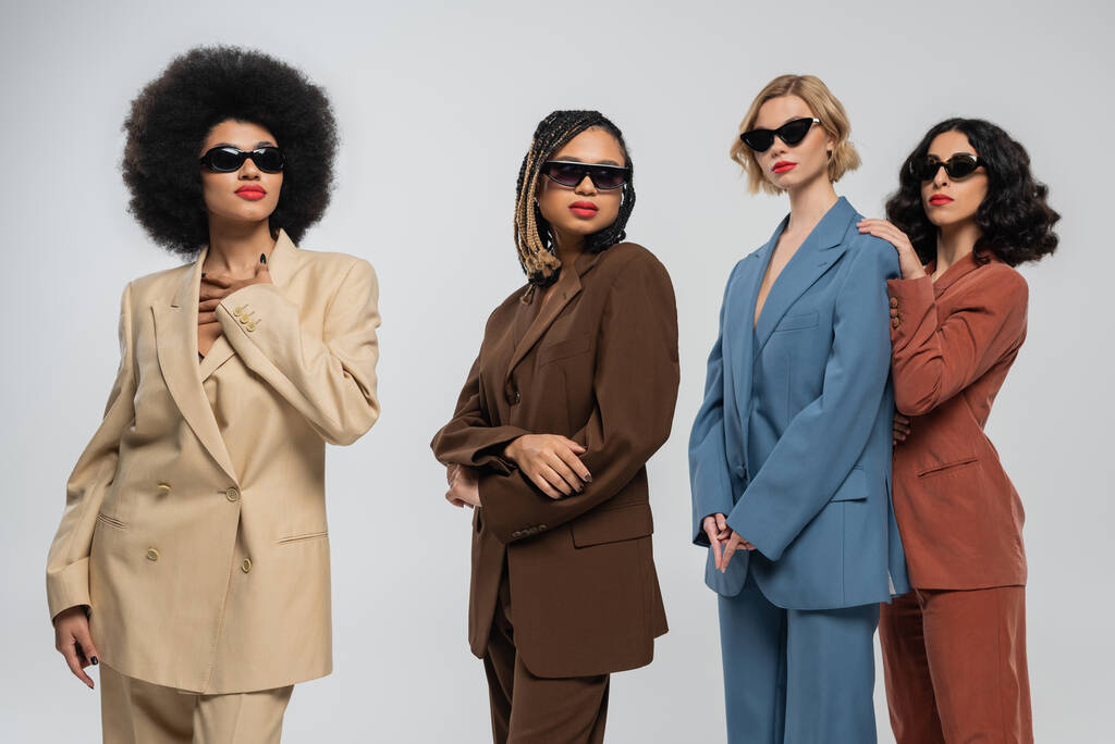 groep van trendy multiraciale vriendinnen in donkere zonnebrillen en veelkleurige pakken op grijs, mode - Foto, afbeelding