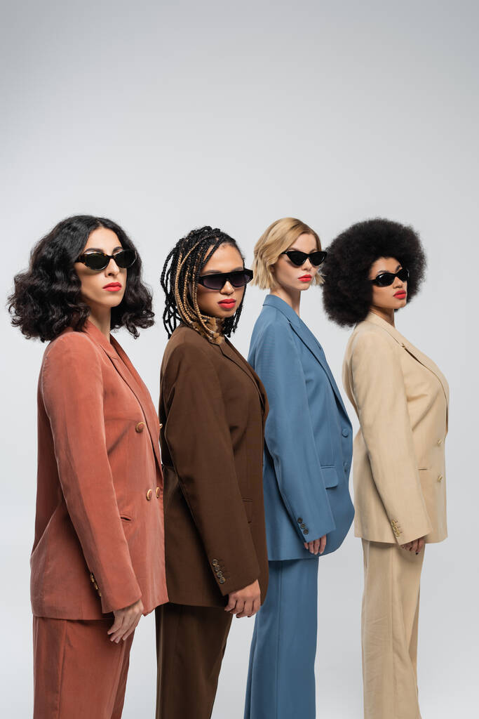 schoonheid in verscheidenheid, multiraciale vrouwen in zonnebrillen en kleurrijke pakken kijken naar camera op grijs - Foto, afbeelding