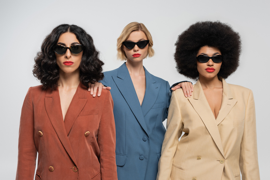 разнообразие и стиль, мультикультурные женщины в модных красочных костюмах и темных солнцезащитных очках на сером - Фото, изображение