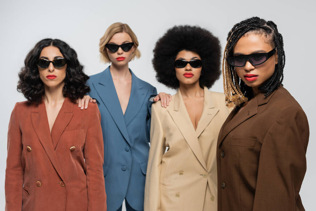 ryhmä trendikkäitä monirotuisia naispuolisia ystäviä tummissa aurinkolaseissa ja värikkäissä puvuissa harmaalla taustalla - Valokuva, kuva