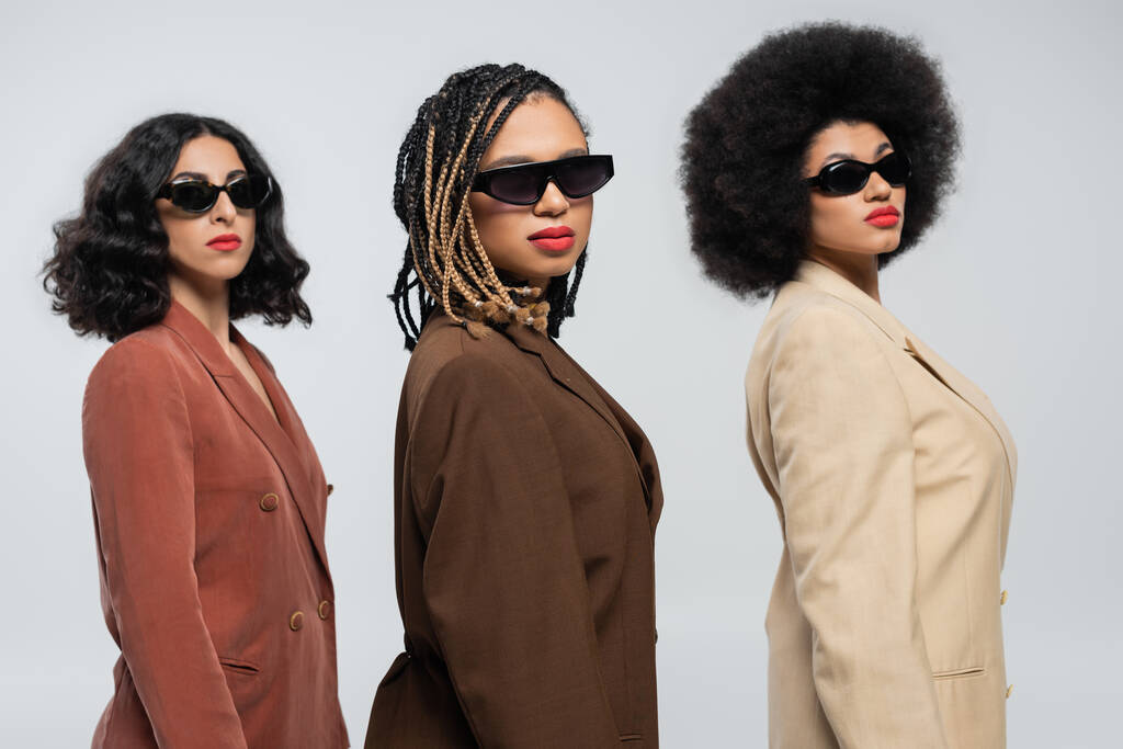 Selbstbewusster Stil, diverse multiethnische Freundinnen in Anzügen und Sonnenbrillen blicken in die Kamera auf grau - Foto, Bild