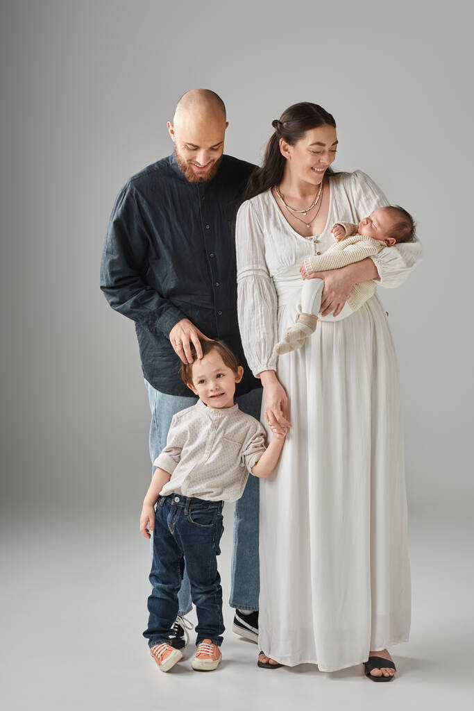 vertikale Aufnahme moderner glücklicher Eltern, die mit ihren kleinen Kindern auf grauem Hintergrund posieren - Foto, Bild