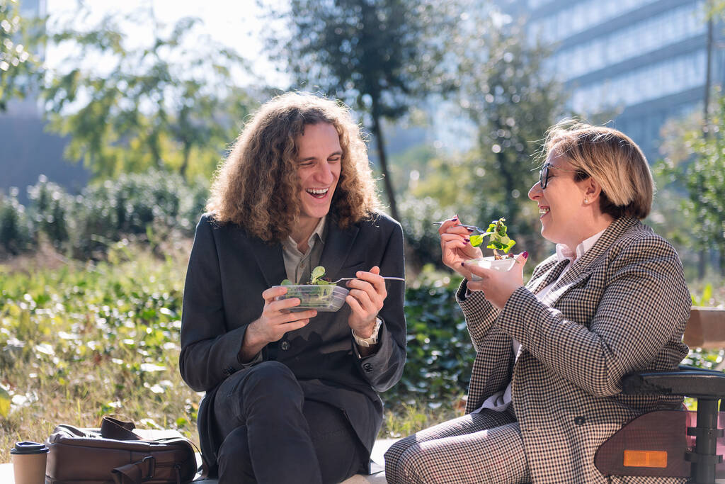 junger Geschäftsmann und Frau im Rollstuhl lächeln glücklich, während sie in einer Arbeitspause draußen einen Salat essen, Konzept der Vielfalt und des urbanen Lebensstils, Kopierraum für Text - Foto, Bild