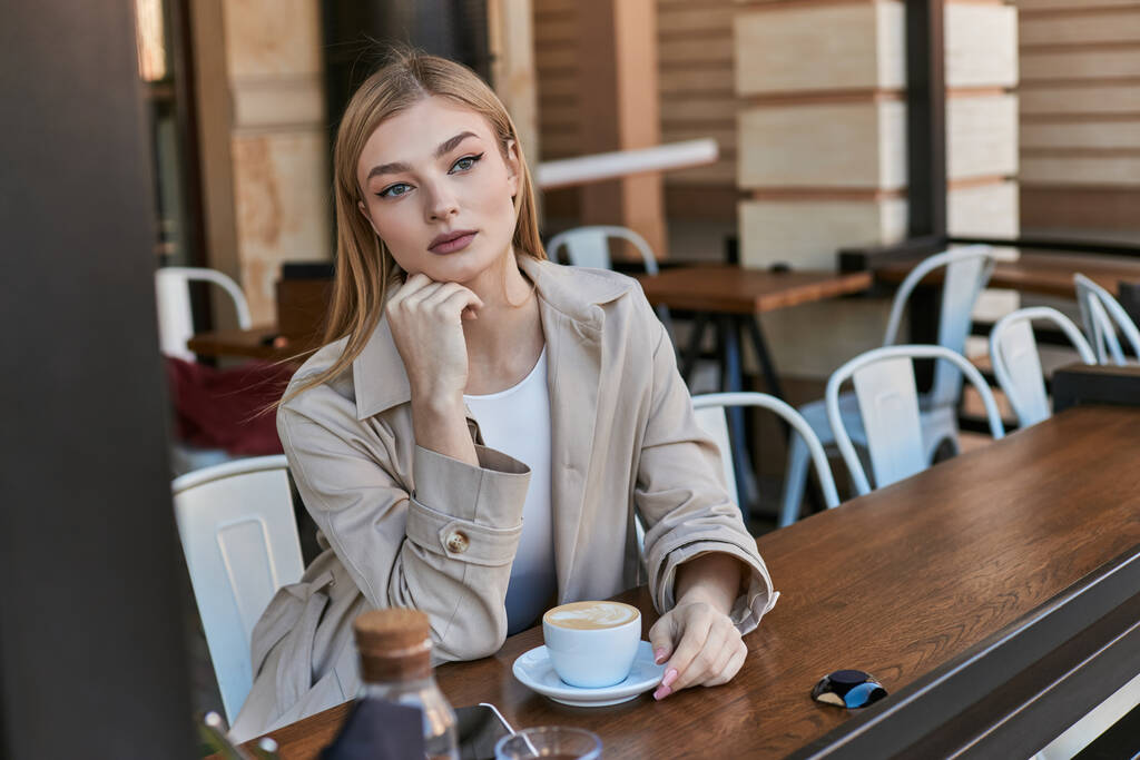 verträumte blonde Frau im Trenchcoat genießt ihre Tasse Cappuccino im Freien im Café, Wochenendstimmung - Foto, Bild