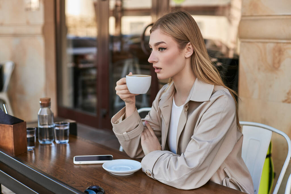 ονειρική νεαρή γυναίκα σε καμπαρντίνα απολαμβάνοντας φλιτζάνι καπουτσίνο, ενώ κάθεται κοντά στο smartphone στο καφέ - Φωτογραφία, εικόνα