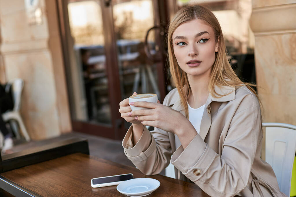красивая молодая женщина в плаще наслаждается чашкой капучино, сидя рядом со смартфоном в кафе - Фото, изображение