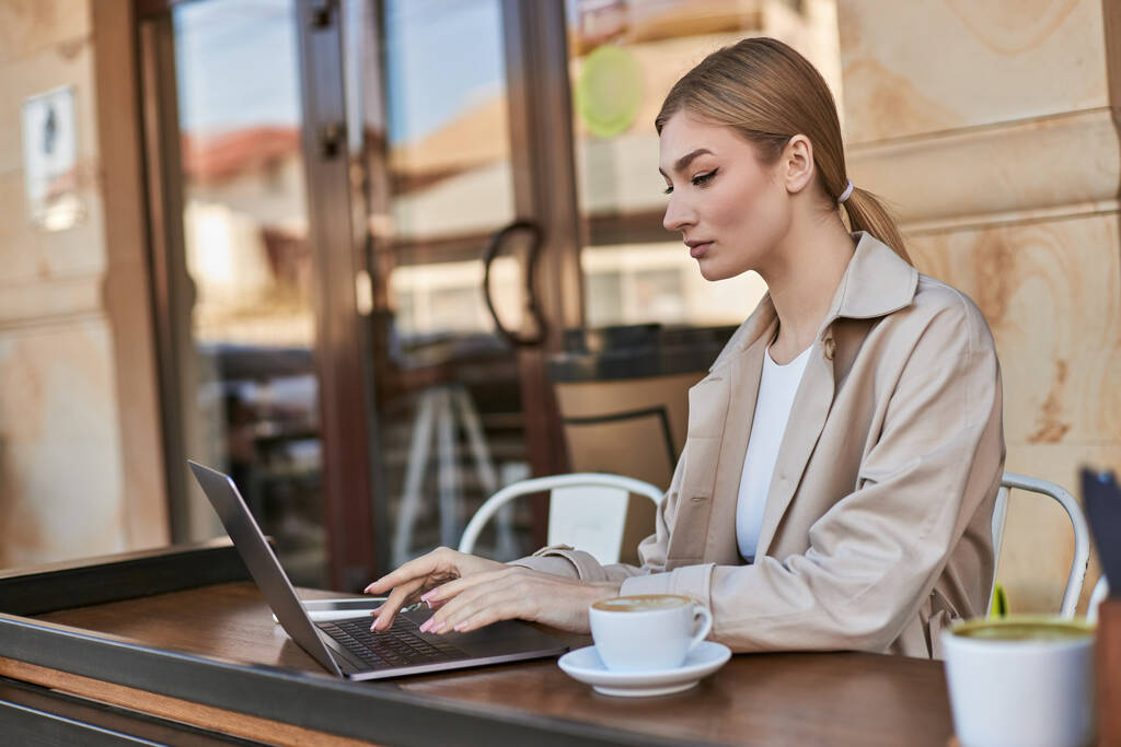νεαρή ξανθιά γυναίκα δακτυλογράφηση σε φορητό υπολογιστή, ενώ εργάζονται εξ αποστάσεως κοντά φλιτζάνι καφέ σε εξωτερικούς χώρους καφέ - Φωτογραφία, εικόνα