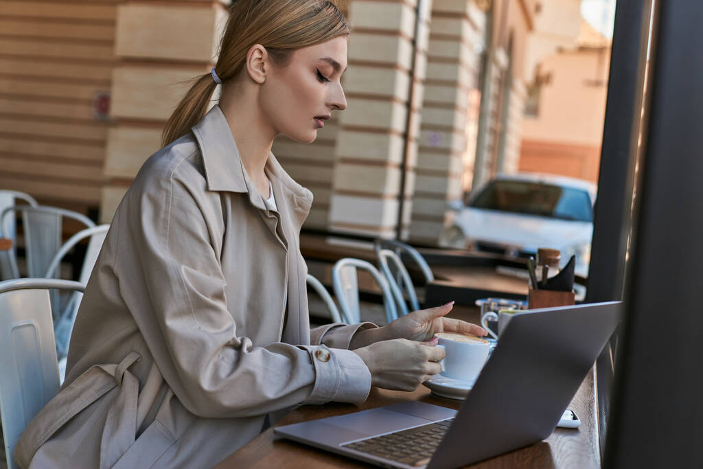 young blonde woman taking cup of coffee near laptop on table in cafe outdoors, remote work - Photo, Image