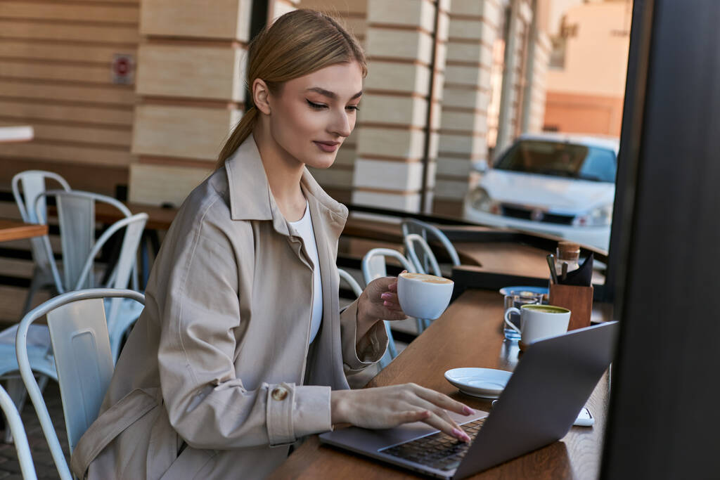 mladá blondýnka drží šálek kávy a pomocí notebooku na stole v kavárně venku, vzdálená práce - Fotografie, Obrázek