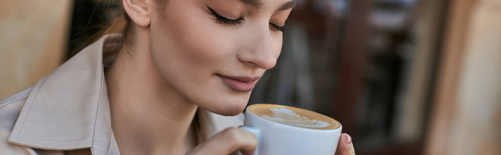spandoek van mooie blonde vrouw in trench jas genieten van haar kopje cappuccino in cafe - Foto, afbeelding