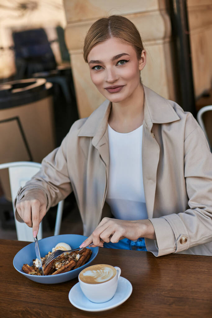 щаслива молода жінка в траншеї пальто насолоджується її бельгійські вафлі з морозивом поруч з чашкою кави - Фото, зображення