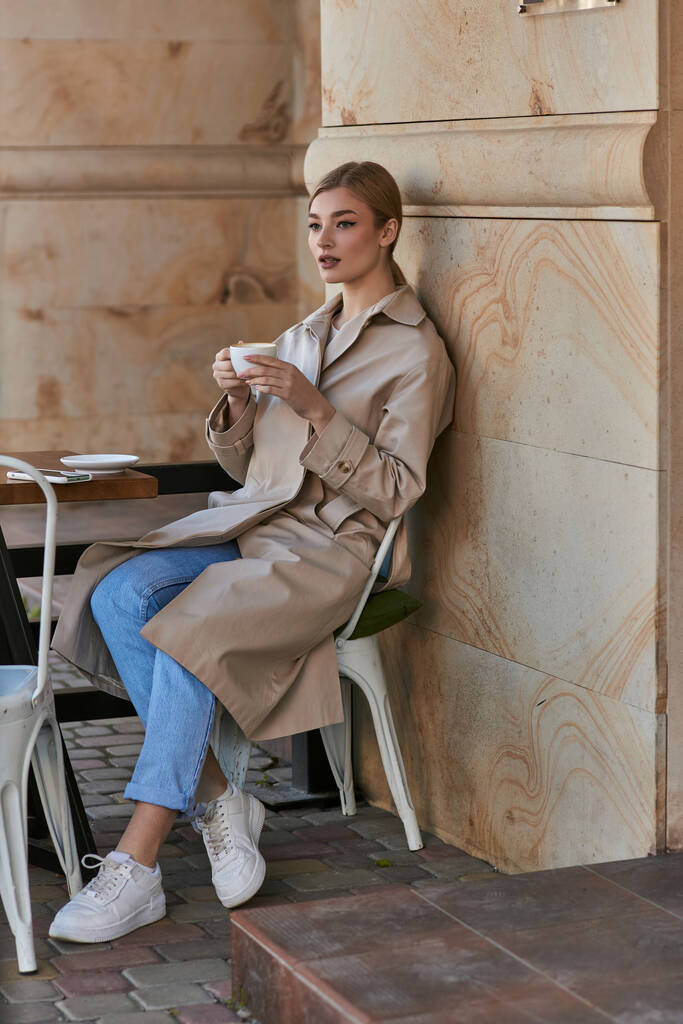 schöne junge Frau in stilvollem Trenchcoat sitzt und hält eine Tasse Kaffee in einem modernen Café - Foto, Bild