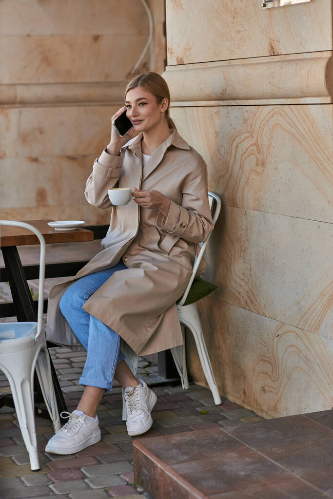 glückliche junge Frau im schicken Trenchcoat, die mit dem Smartphone spricht und im Café eine Tasse Kaffee hält - Foto, Bild