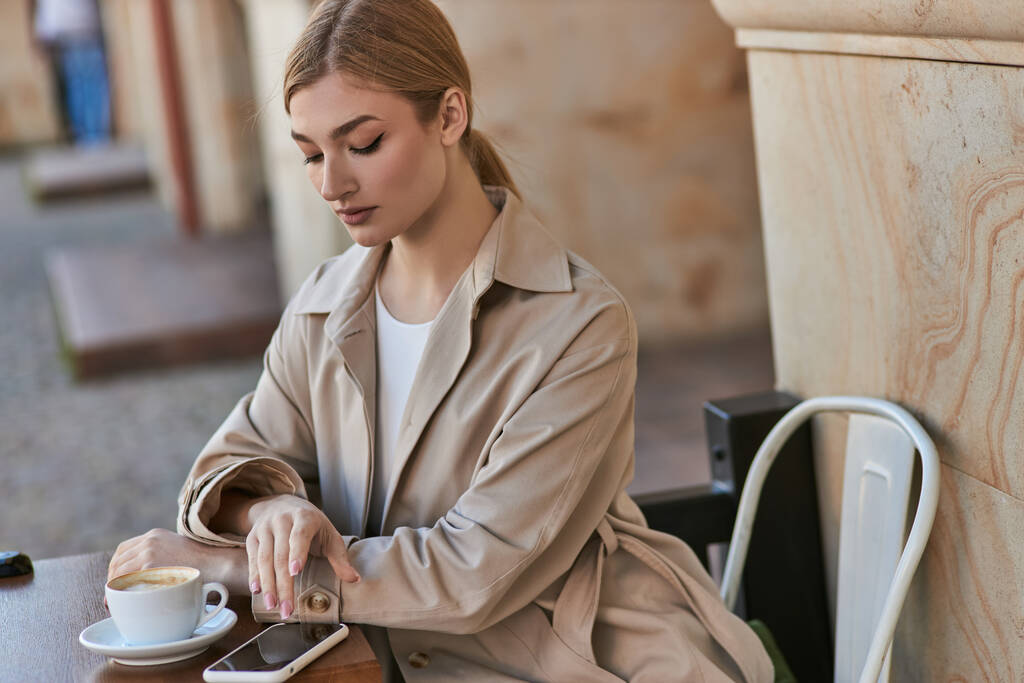 ξανθιά νεαρή γυναίκα σε κομψό καμπαρντίνα χρησιμοποιώντας smartphone κοντά φλιτζάνι καφέ στο τραπέζι στο καφέ - Φωτογραφία, εικόνα