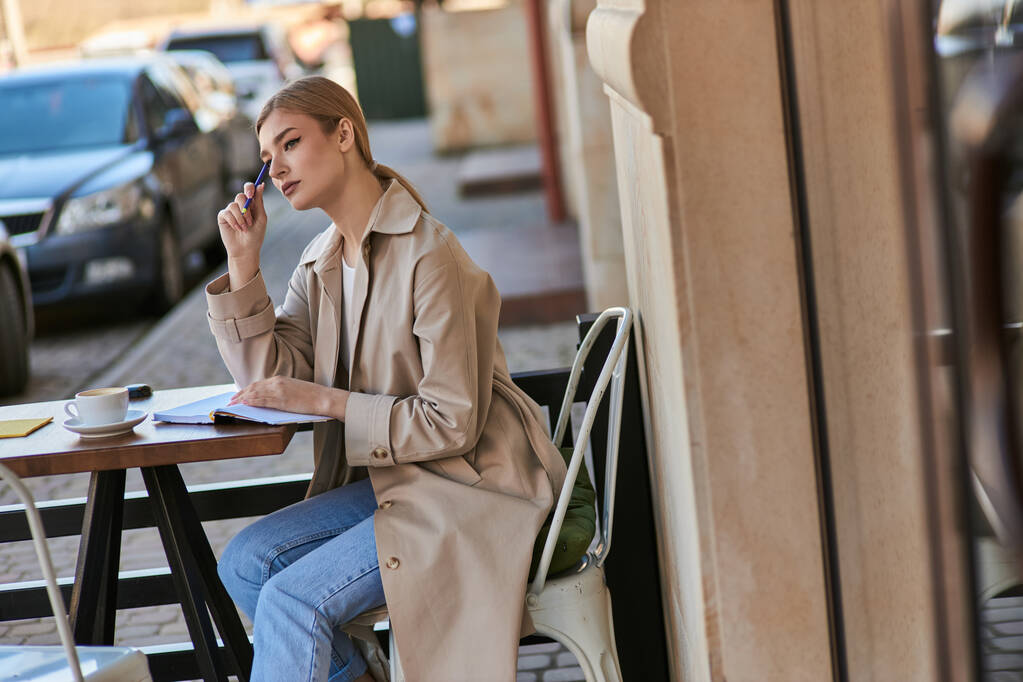 Trençkotlu düşünceli bir kadın bir fincan kahve ve elinde kalemle masada oturuyor, günlük yazıyor. - Fotoğraf, Görsel