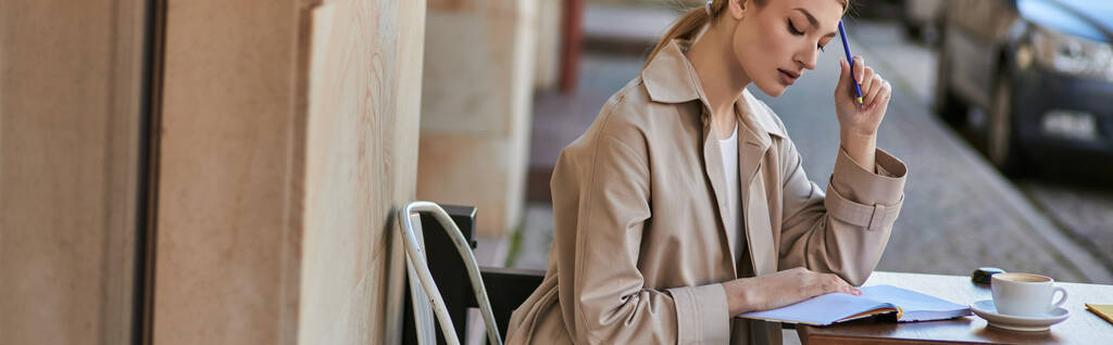 женщина в плаще сидит рядом с чашкой кофе и держит ручку возле блокнота, пишет баннер дневника - Фото, изображение