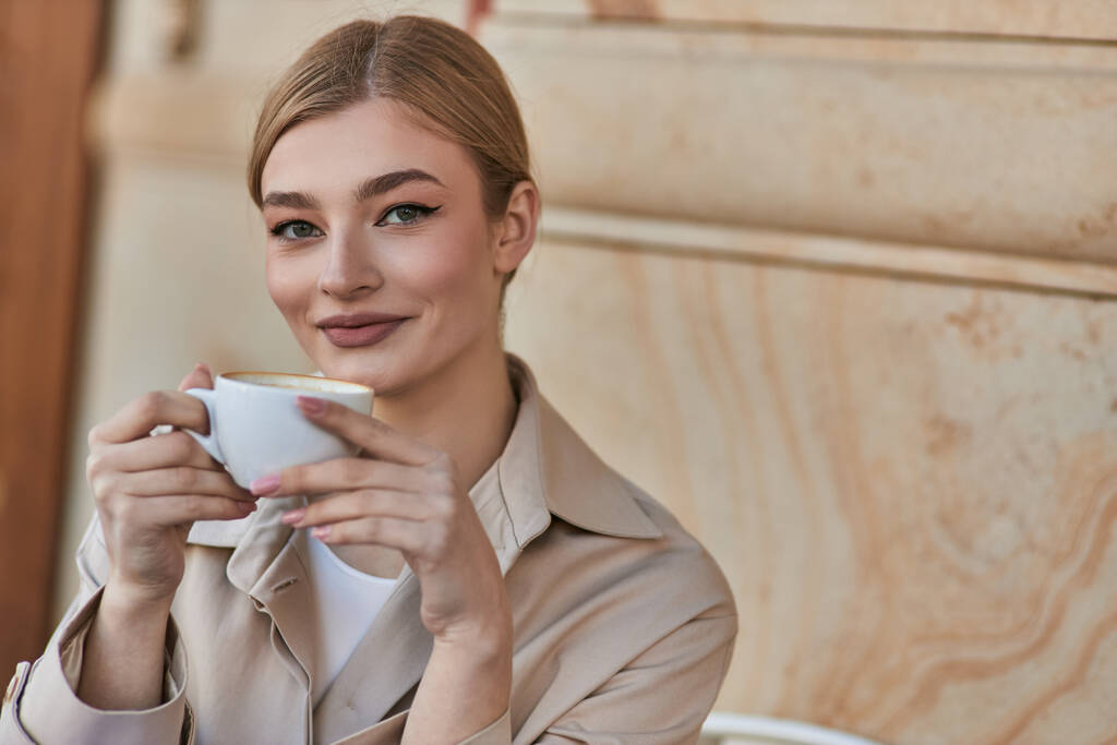 χαρούμενη ξανθιά γυναίκα σε κομψό καμπαρντίνα κρατώντας φλιτζάνι καφέ, ενώ κάθεται στο καφέ, ποτό - Φωτογραφία, εικόνα