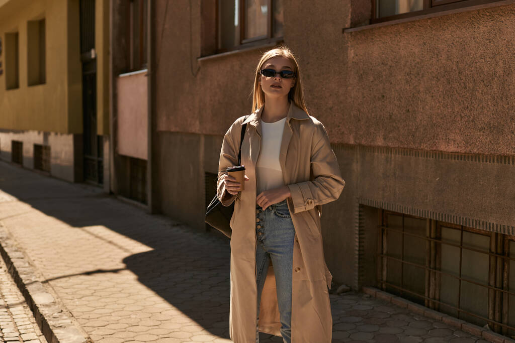 elegantní žena ve stylovém zákopu kabát drží odnést kávu, zatímco stojí na ulici ve slunečný den - Fotografie, Obrázek