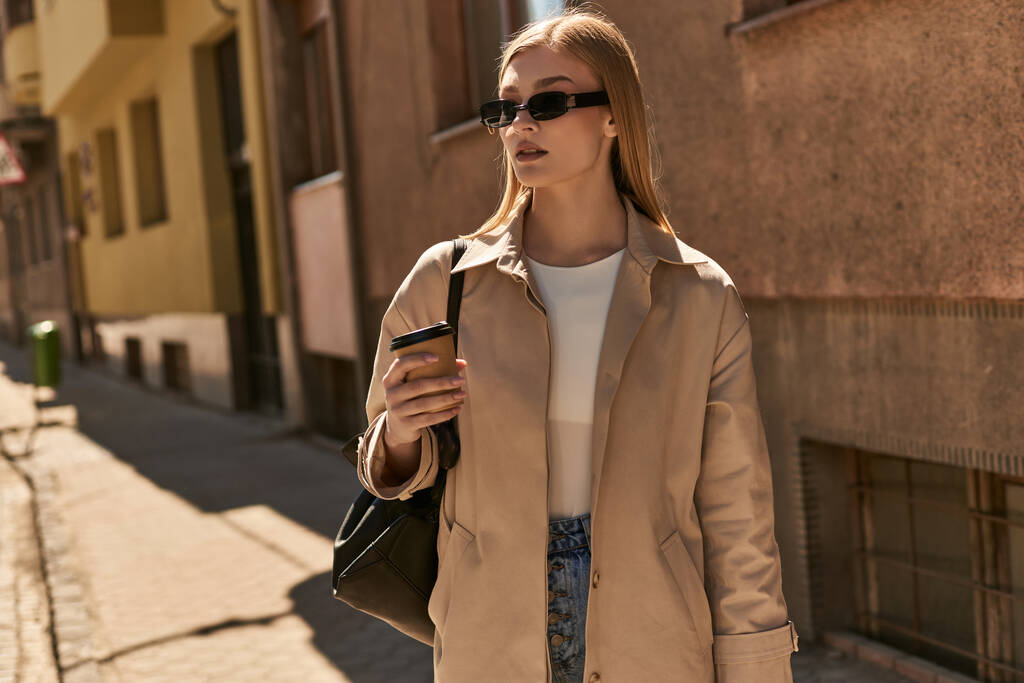 hezká žena ve stylovém zákopu kabát drží odnést kávu, zatímco stojí na ulici ve slunečný den - Fotografie, Obrázek