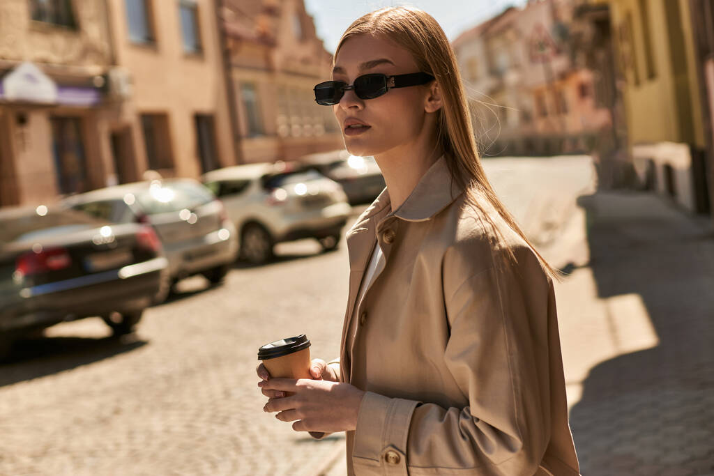 blond vrouw in stijlvolle trench jas houden van koffie te gaan tijdens het lopen op straat in zonnige dag - Foto, afbeelding