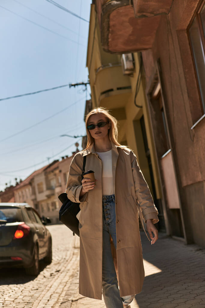 блондинка молода жінка в траншеї пальто і сонцезахисні окуляри тримає каву, щоб йти і ходити по вулиці - Фото, зображення