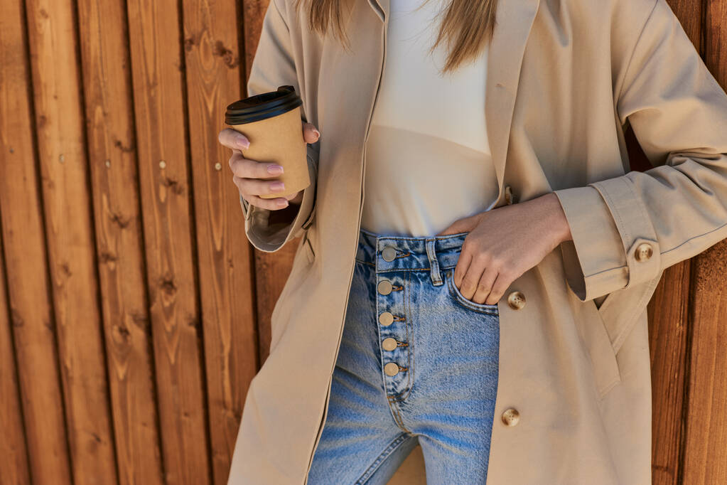 обрізана жінка в стильному траншеї пальто і сонцезахисні окуляри, що тримають каву, щоб піти і стояти біля паркану - Фото, зображення
