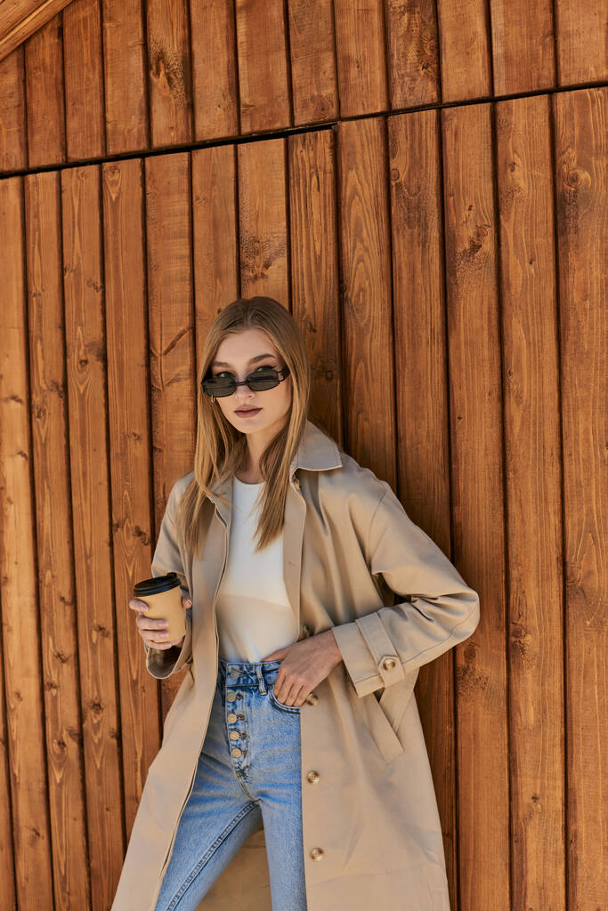 mujer rubia en gabardina y gafas de sol sosteniendo café para ir y de pie con la mano en el bolsillo - Foto, Imagen