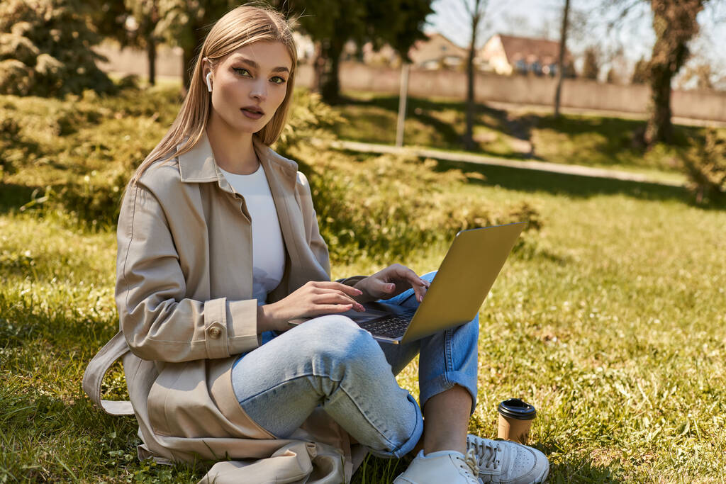 Blondine in Kopfhörer und Trenchcoat mit Laptop im Gras neben Pappbecher - Foto, Bild