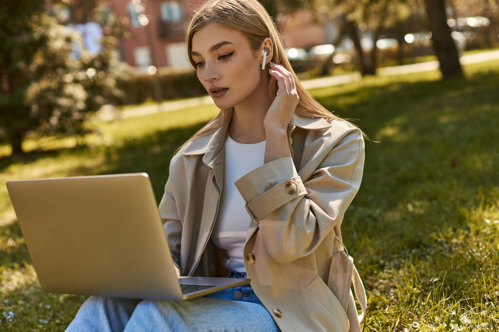 blonde jeune femme dans les écouteurs sans fil et trench coat à l'aide d'un ordinateur portable tout en étant assis sur l'herbe - Photo, image