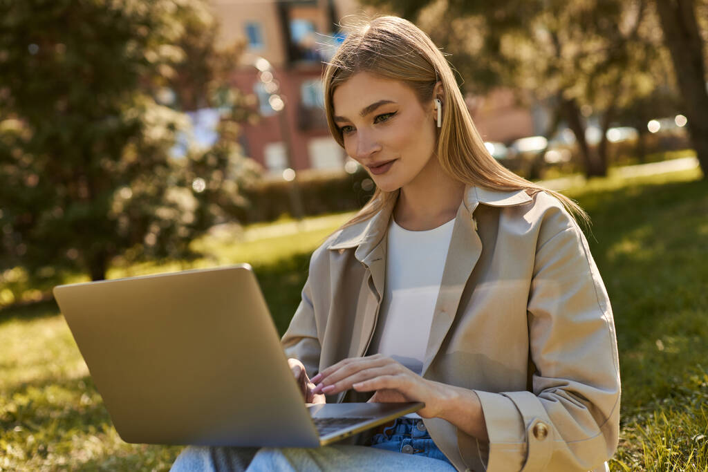 aantrekkelijke jonge vrouw in draadloze oortelefoons en trench jas met behulp van laptop tijdens het zitten op gras - Foto, afbeelding