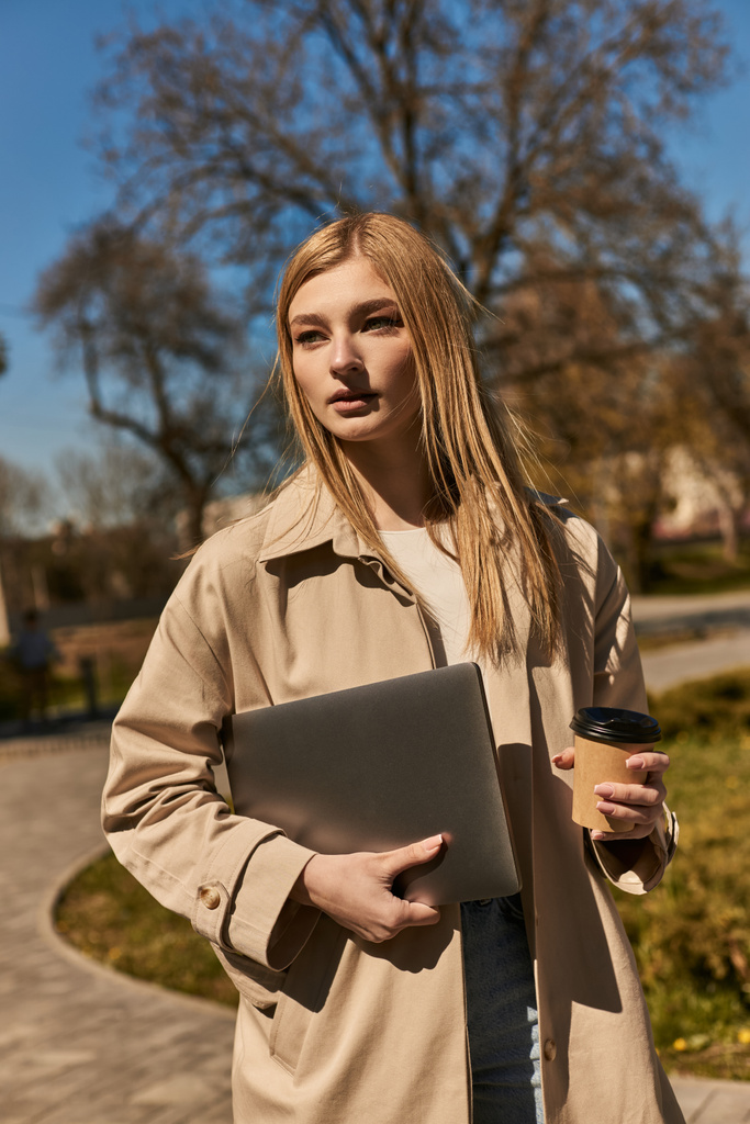 mladá blondýnka ve stylové trenč kabát drží papírový šálek s kávou a notebookem v parku - Fotografie, Obrázek