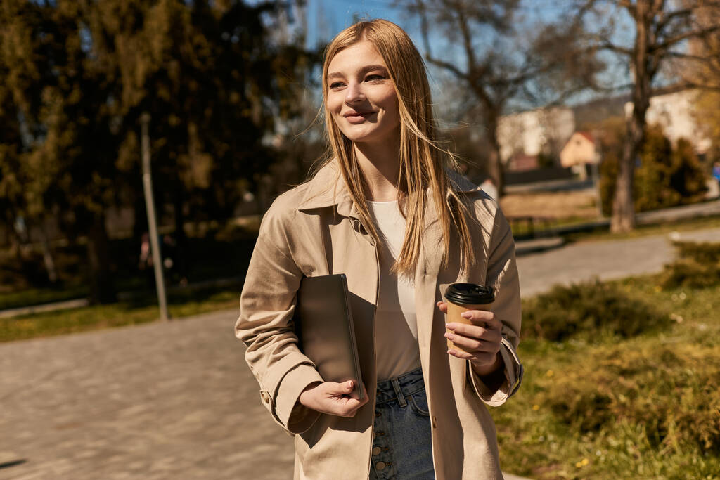 щаслива блондинка в стильному траншеї пальто тримає паперову чашку з кавою і ноутбуком в парку - Фото, зображення