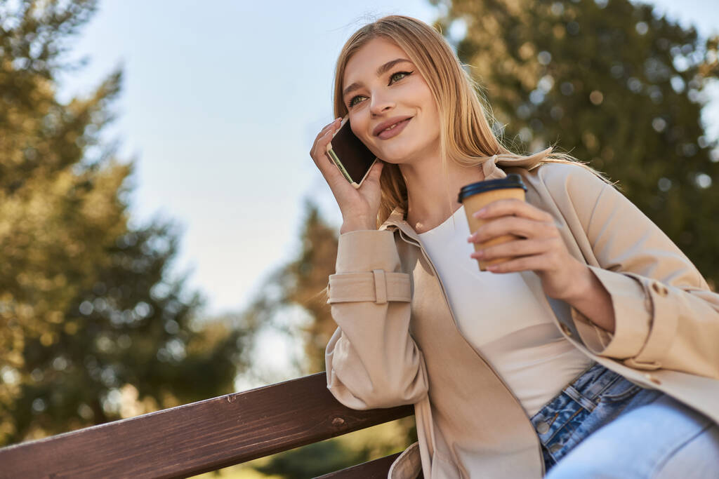 mujer feliz en gabardina sosteniendo café para llevar y hablando en el teléfono inteligente mientras está sentado en el banco - Foto, imagen