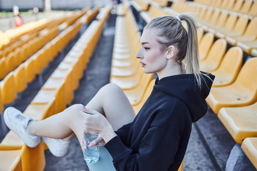 Blonde Sportlerin mit Pferdeschwanz hält Wasserflasche in der Hand und sitzt nach dem Training auf dem Stadionstuhl - Foto, Bild