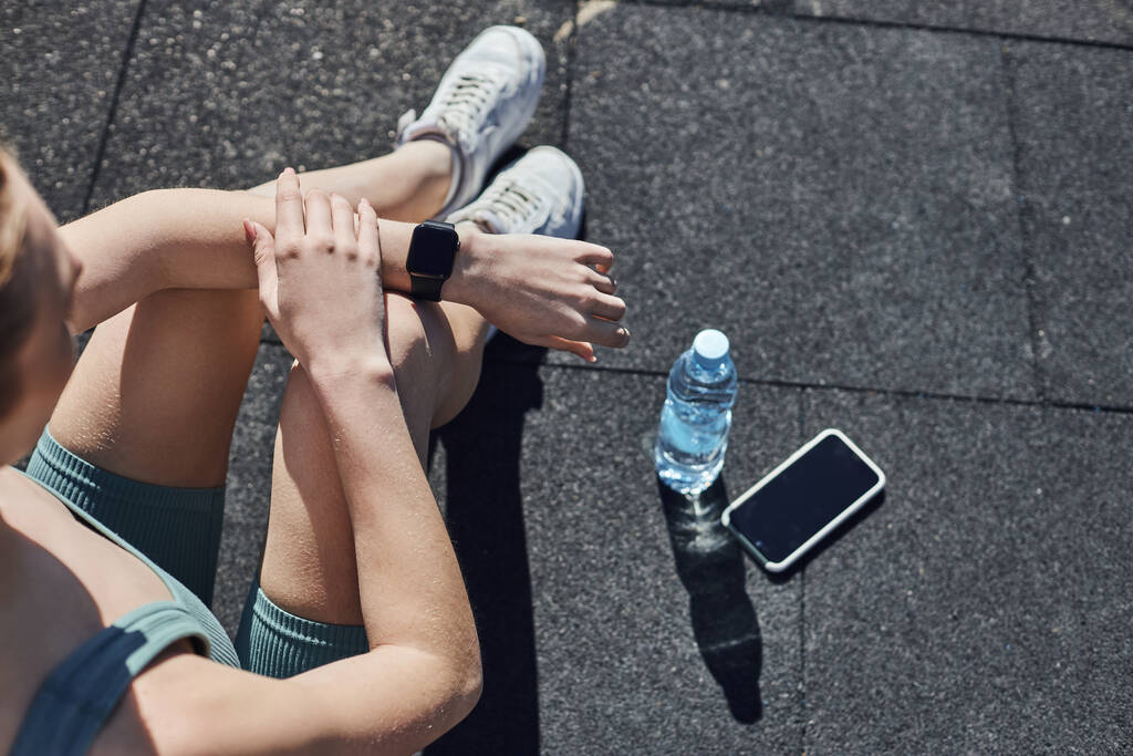 κορυφαία άποψη της ταιριάζει γυναίκα σε activewear έλεγχο fitness tracker δίπλα στο smartphone και μπουκάλι νερό - Φωτογραφία, εικόνα