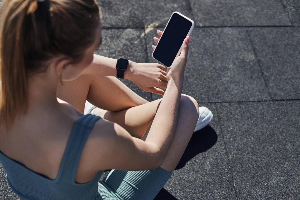 κορυφαία άποψη της ταιριάζει γυναίκα σε activewear με tracker γυμναστήριο στον καρπό χρησιμοποιώντας smartphone μετά την προπόνηση - Φωτογραφία, εικόνα