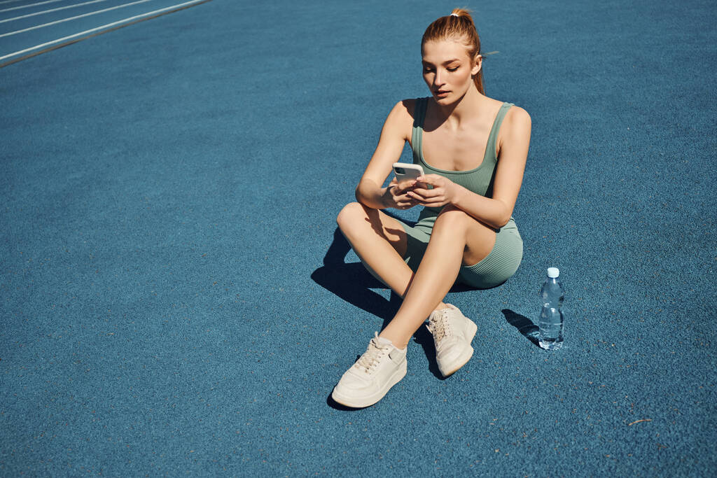 Sportos nő aktív viseletben ül egy üveg víz mellett és mobiltelefont használ edzés után - Fotó, kép