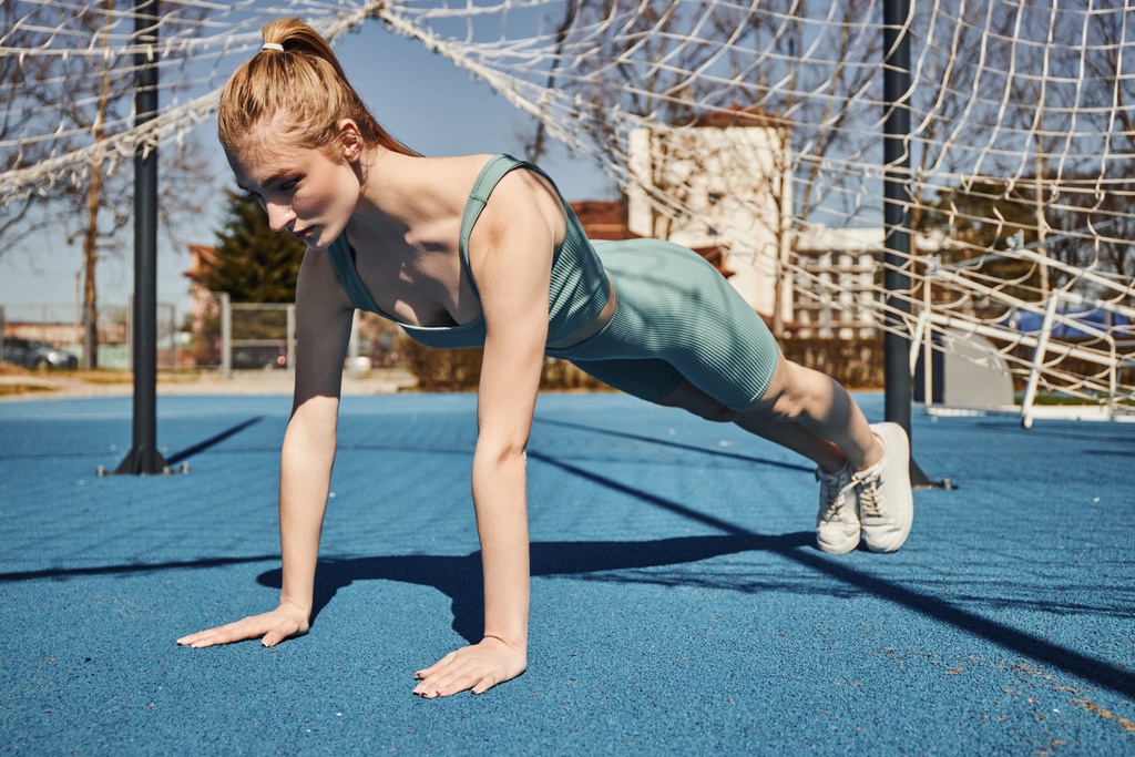blonde young sportswoman with ponytail exercising in activewear near net outdoors, doing press ups - Photo, Image