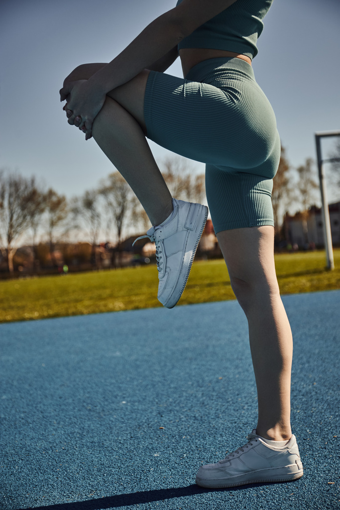 частичный взгляд на гибкую спортсменку, тренирующуюся в плотно активной одежде на открытом воздухе, растягивающую ногу - Фото, изображение