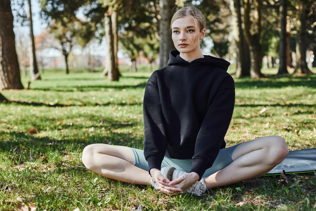 sportieve jonge vrouw met blond haar en sportkleding zittend op mat en mediterend in het park - Foto, afbeelding