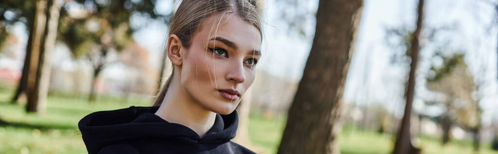 portrait of sporty young woman in black hoodie looking away in green park, nature and sport banner - Photo, Image