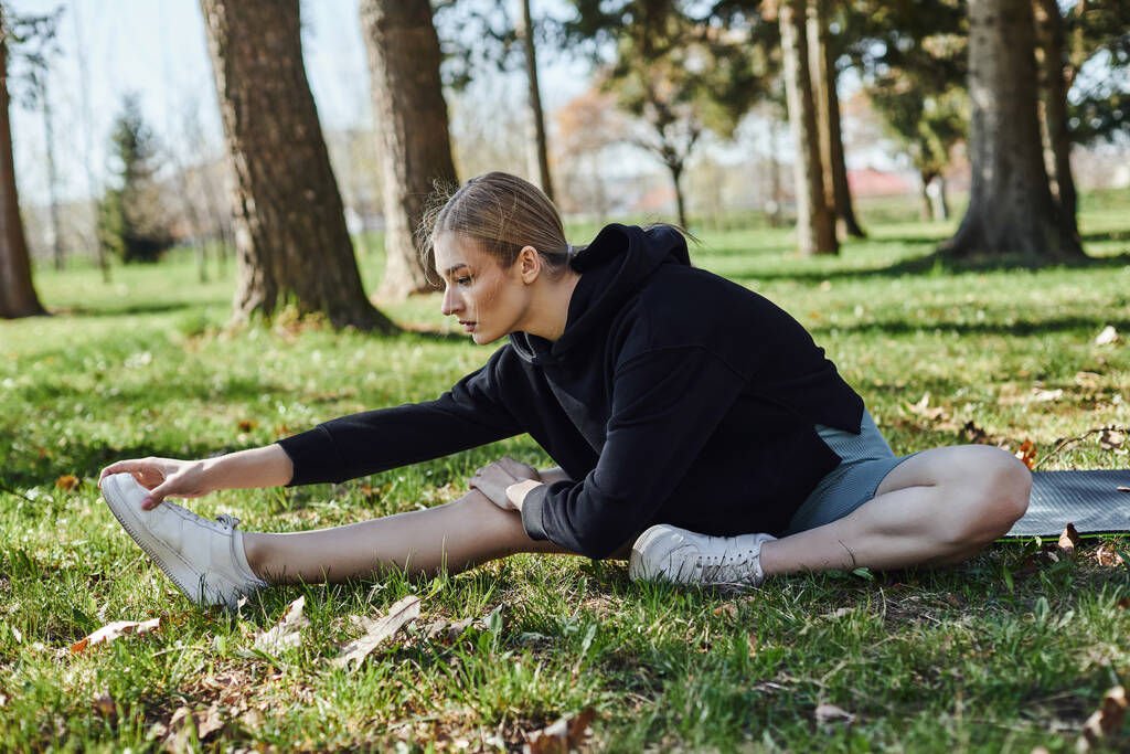 fitte junge Frau mit blonden Haaren und Sportbekleidung beim Dehnen im Park auf einer Fitnessmatte sitzend - Foto, Bild