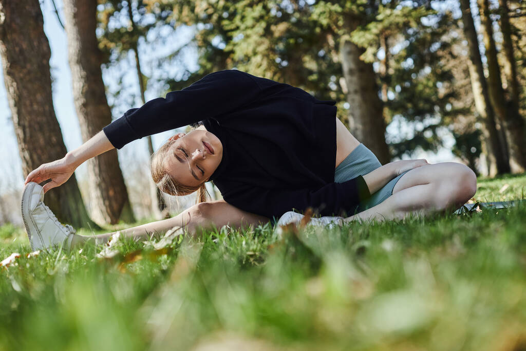 sporty young woman with blonde hair and sportswear stretching while sitting on green grass in park - Photo, Image
