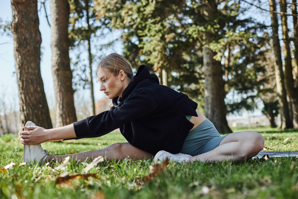 fitte junge Frau mit blonden Haaren und Sportbekleidung, die das Bein streckt, während sie im Park auf einer Fitnessmatte sitzt - Foto, Bild