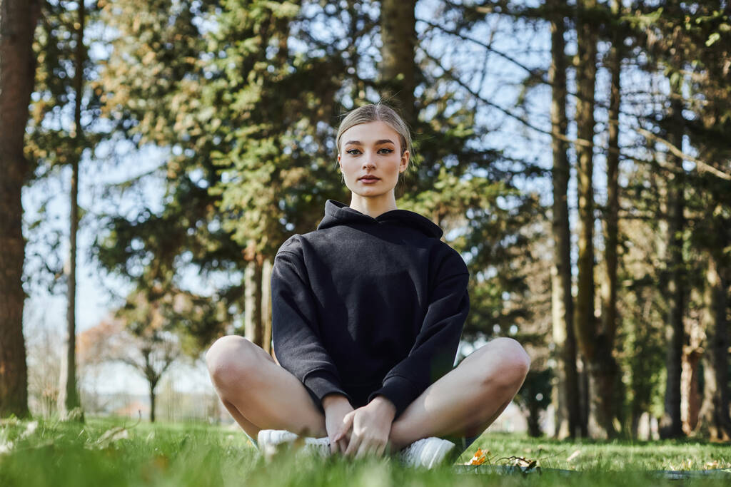 mujer bonita y en forma con el pelo rubio y ropa deportiva sentado en la estera mientras medita en el parque - Foto, imagen